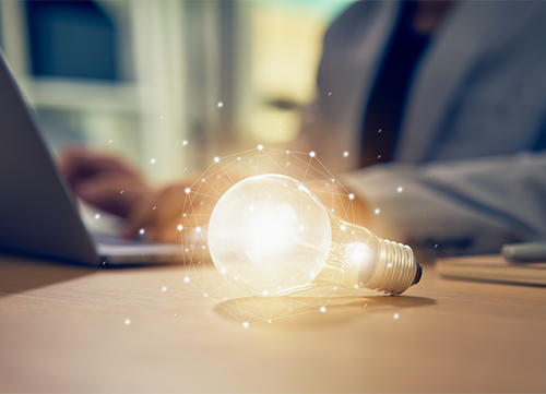 A glowing lightbulb on a table in front of a man working on a laptop.